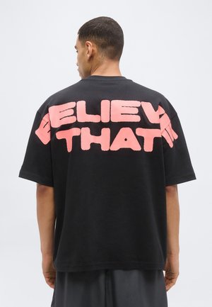 Uomo in piedi con la schiena rivolta verso la fotocamera, indossa una T-shirt nera oversize con grandi lettere rosa che parzialmente leggono "BELIEV THAT" sulle spalle.