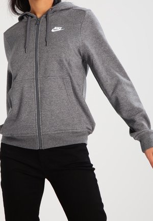 Personne portant un sweat à capuche gris Nike zippé avec une poche avant et un pantalon noir sur un fond blanc uni.