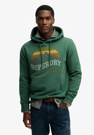 Giovane uomo che indossa una felpa verde Superdry sopra una camicia a quadri, con jeans blu e una cintura intrecciata marrone, in piedi con una mano in tasca.