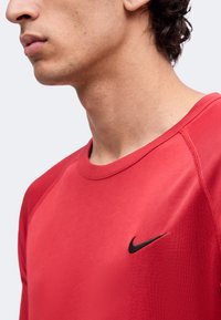 Νεαρό άτομο που φοράει κόκκινο πουκάμισο Nike με μαύρο λογότυπο swoosh, που εμφανίζει τον λαιμό, το πηγούνι, το αυτί και την ανώτερη περιοχή του στήθους.