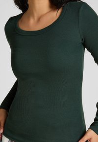 Geribbelde langekomshirt in donkergroen. Heeft een ronde halslijn en een aansluitend ontwerp, dat een gladde textuur en rekbare stof toont.