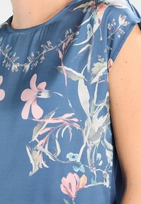 Modrý květinový top s krátkým rukávem vyrobený z hladké, lehké látky s růžovými a zelenými botanickými vzory kolem výstřihu a ramen.