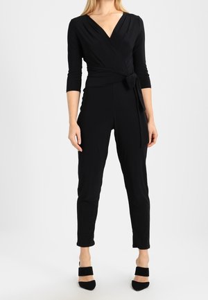 Schwarzer Wickel-Jumpsuit mit langen Ärmeln und V-Ausschnitt, tailliertem Gürtel, eng anliegenden Beinen und glattem Stoff. Kombiniert mit schwarzen High Heels.