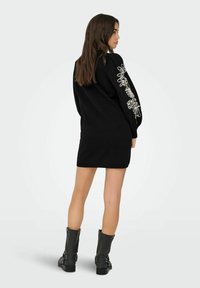 Zwart gebreid jurkje met lange mouwen en witte bloemen borduursels, een relaxte pasvorm en ribgebreide manchetten. Gecombineerd met zwarte enkellaarsjes.