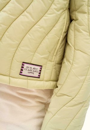 Rękaw i tył jasnooliwkowej pikowanej kurtki z prostokątną naszywką z napisem "ZA CHWILĘ ZDARY SIĘ MAGIA."