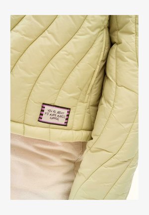 Rękaw i tył jasnooliwkowej pikowanej kurtki z prostokątną naszywką z napisem "ZA CHWILĘ ZDARY SIĘ MAGIA."