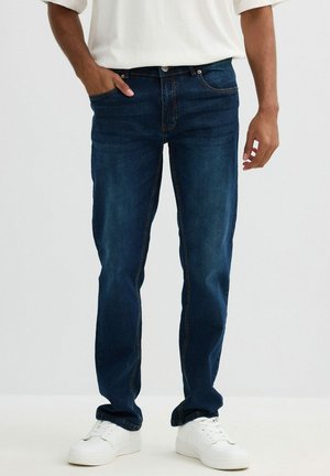 Los pantalones vaqueros de mezclilla azul oscuro tienen un corte recto, cuatro bolsillos y detalles de costura sutiles. Combinados con zapatillas blancas.