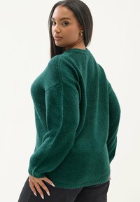 Zielony dziany sweter o luźnym kroju, z dużymi rękawami i teksturalnym wykończeniem. Materiał wydaje się miękki i przytulny.