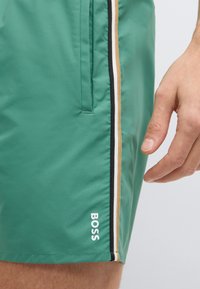 Zelené plavecké šortky z hladkej látky s bočným pruhovaným dizajnom v čiernej, bielej a hnedej farbe, s logom na leme.