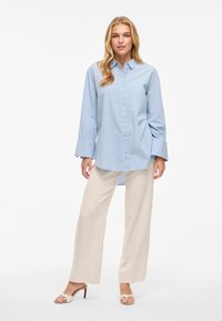 Gaiši zila krekla krekls ar garām piedurknēm, ar taisnu piegriezumu un maigu tekstūru. Saskaņots ar bēšām platām biksēm un papēžu sandālēm.