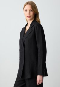 Czarna marynarka wykonana z teksturowanej tkaniny, z jednorzędowym zapięciem, dwoma guzikami i dopasowanym krojem. Stylizowana z naszyjnikiem.