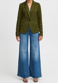 Grüner strukturierter Blazer mit tailliertem Schnitt, zwei Fronttaschen und einem einzigen Knopf. Kombiniert mit weiten blauen Jeans und schwarzen Absätzen.