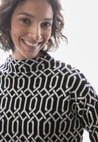 Personne souriante aux cheveux courts et bouclés portant un pull à col montant noir et blanc à motif, sur un fond blanc.