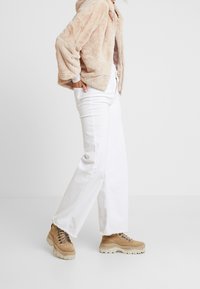 Beżowa kurtka z futerkiem, białe jeansy z wysokim stanem oraz brązowe buty na masywnej podeszwie. Kurtka wyposażona w kaptur i zapięcie na zamek, jeansy o prostych nogawkach.