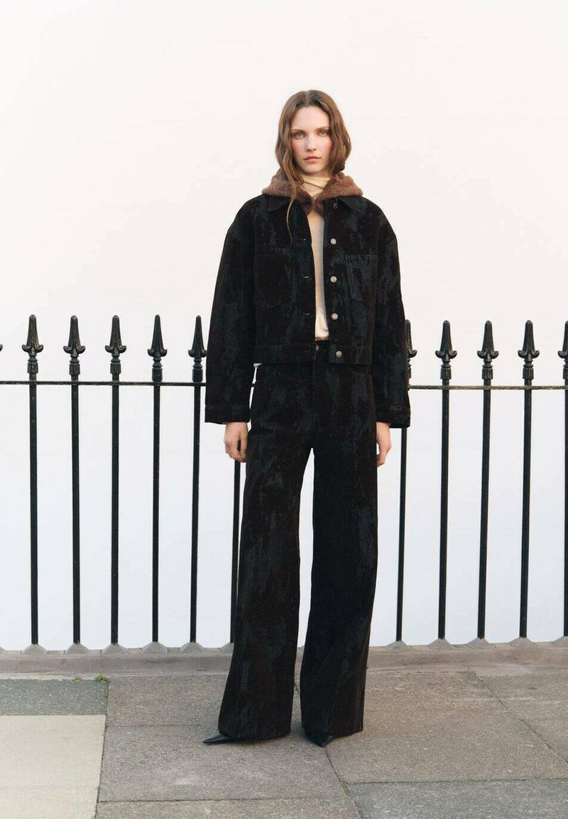 Jachetă din catifea neagră cu guler din blană maro, asortată cu pantaloni evazați negri, cu talie înaltă. Ambele au un finisaj texturat și detalii cu nasturi.