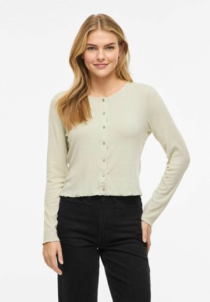 Jeune femme aux longs cheveux blonds portant un cardigan beige clair boutonné et un pantalon noir, debout, une main sur la hanche.