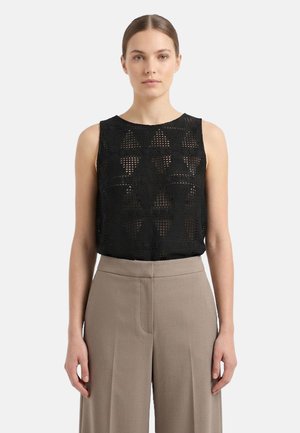 Donna che indossa un top nero senza maniche all'uncinetto con motivo geometrico e pantaloni beige a vita alta a gamba larga, in piedi davanti a uno sfondo semplice.
