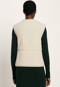 Gepolsterte, ärmellose Weste in hellbeige mit glatter Textur. Verfügt über einen hohen Kragen und eine Naht im Rücken, auf einer Person präsentiert.