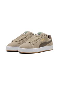 Beige suede sneakers with white rubber sole, brown accents, and green tags featuring the Puma logo. Thick laces and classic low-top design.