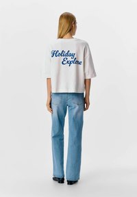 Camiseta corta blanca con texto azul en cursiva "Holiday Explore" en la parte posterior, combinada con jeans azul claro y zapatos negros.