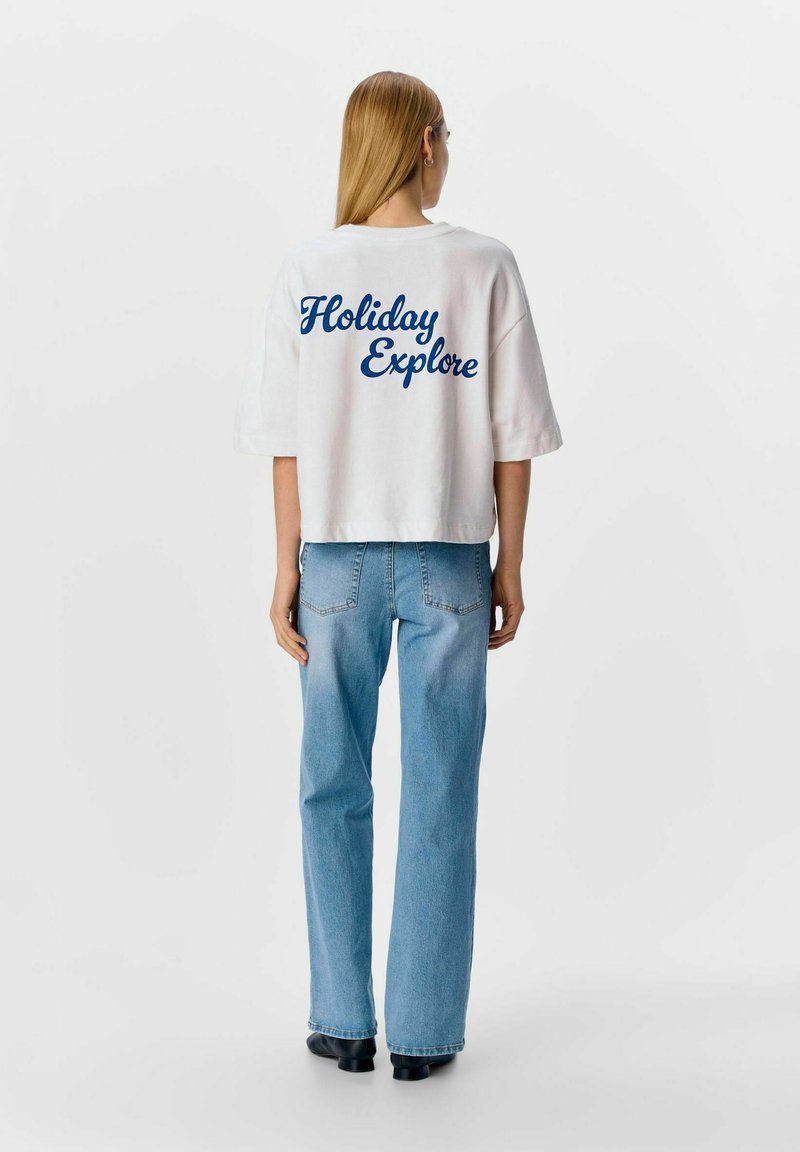 Camiseta corta blanca con texto azul en cursiva "Holiday Explore" en la parte posterior, combinada con jeans azul claro y zapatos negros.