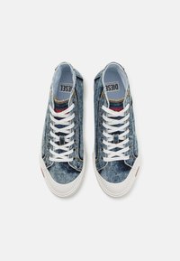 Džinsiniai sportbačiai su šviesiai mėlyna danga, baltais raišteliais ir kontrastinga guma. Prekės ženklas matomas ant liežuvėlio ir šoninių akcentų.