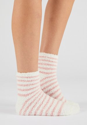 Un par de piernas con calcetines peludos a rayas blancas y rosas sobre un fondo gris claro liso.