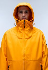 Světlá žlutá nepromokavá bunda s vysokým límcem, nastavitelnou kapucí, zipem vpředu a bočními kapsami. Hladká textura s akcentovaným prošíváním.