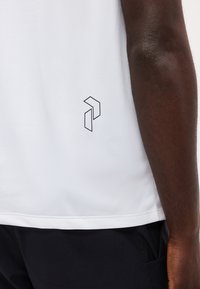 Gros plan d'un bras à la peau foncée à côté de la moitié inférieure d'une chemise blanche avec un petit logo géométrique noir au-dessus de l'ourlet.