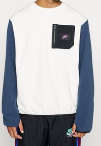 Vit fleece-tröja med marinblå ärmar, försedd med en svart dragkedjeficka och en lila Nike-logotyp på bröstet.