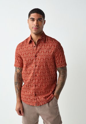 Homme avec des tatouages portant une chemise rouge à motifs à manches courtes et un pantalon beige, debout devant un fond blanc uni.