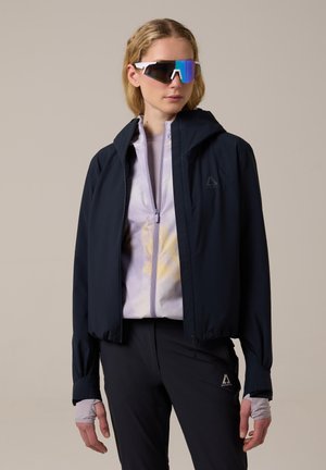 Vrouw die een zwarte windbreaker, lichtpaarse zip-up jas, zwarte broek en grote reflecterende sportzonnebril draagt tegen een effen achtergrond.
