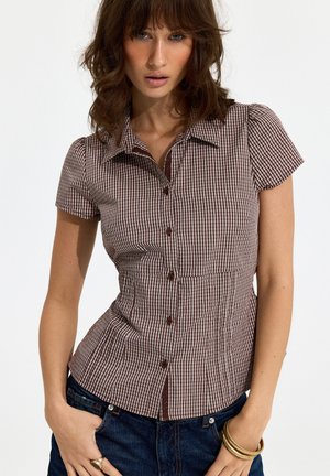 Woman wearing short-sleeve brown and white checkered button-up shirt and dark blue jeans, with hands in pockets and gold bracelets on wrist.