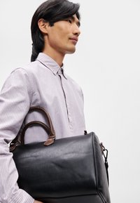 Svart läderväska med en slät yta och rundad form, med bruna läderhandtag och en avtagbar rem. Randig skjorta.