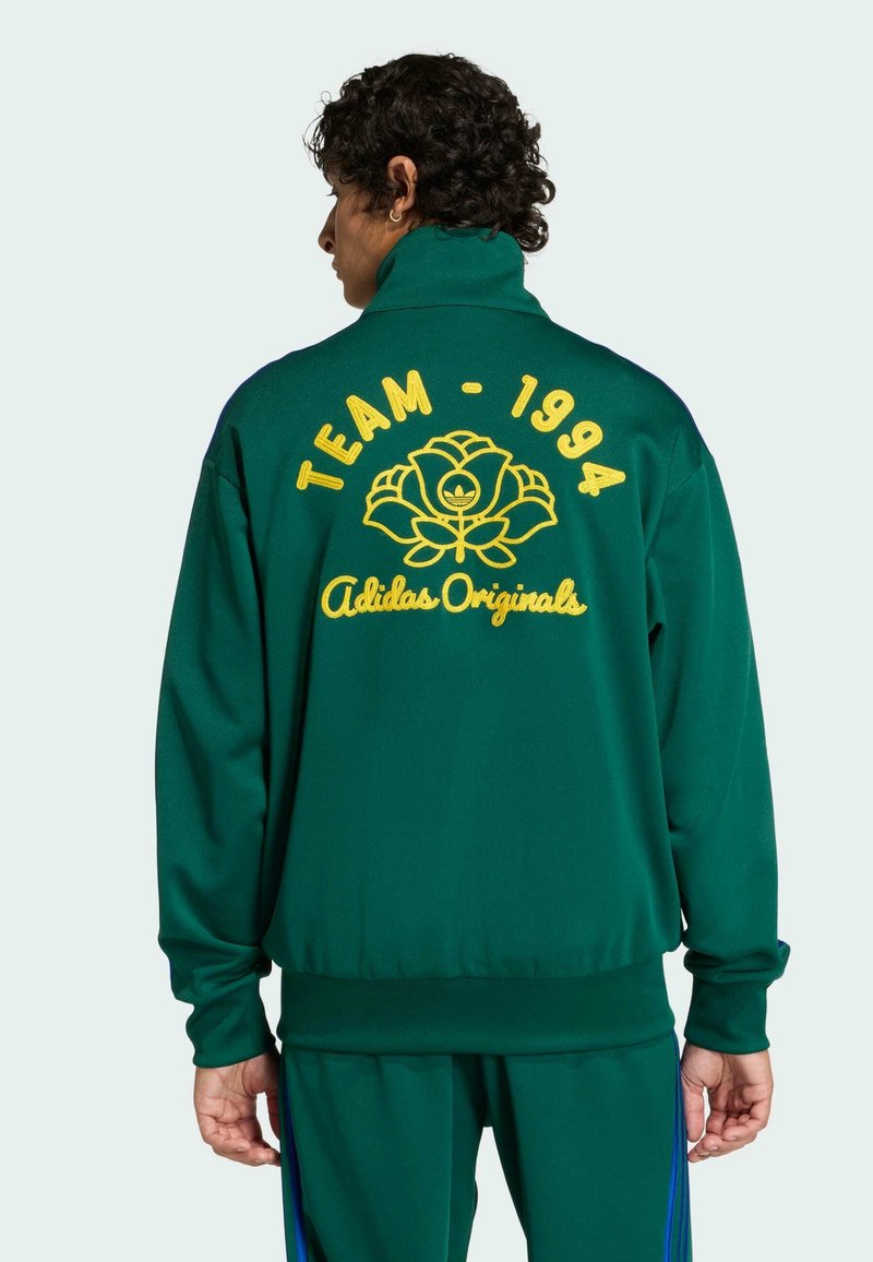 Persona vista di spalle che indossa una giacca sportiva Adidas Originals verde scuro con la scritta gialla "TEAM - 1994" e il logo di un fiore sul retro.