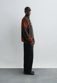 Strikket cardigan i gradient brun og oransje farger, med knapp foran og en mørk skjorte under, parret med svarte bukser og sko.