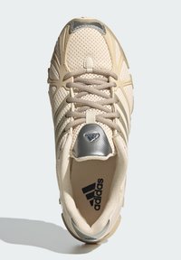 Bežová športová obuv z priedušnej sieťoviny so sivými akcentmi, okrúhlym nosom, šnurovacím dizajnom a výrazným logom na jazyku. Má textúrovanú podrážku a priedušnú vrchnú časť.