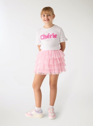 Ragazza giovane sorridente che indossa una maglietta bianca con la scritta "Chérie", gonna in tulle rosa a strati, calzini bianchi alla caviglia e sneakers pastello, in piedi davanti a uno sfondo bianco.