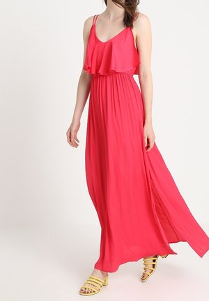 Vrouw draagt een vloeiende, rode, mouwloze maxi-jurk met ruches, gecombineerd met gele sandalen met bandjes en blokhakken.