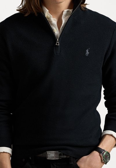 Granatowy sweter dzianinowy z zamkiem przy kołnierzu, z małym haftowanym logo i kontrastowym białym wnętrzem kołnierza.