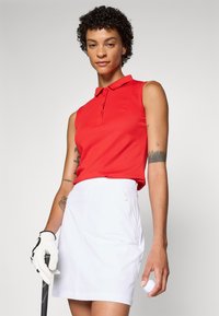 Red sleeveless polo shirt with a collar and a subtle logo, paired with a white fitted golf skirt featuring a zipper detail. Holding a golf ball.