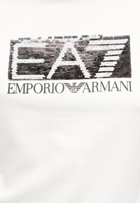 Λογότυπο EA7 με παγιέτες πάνω από το κείμενο Emporio Armani και το έμβλημα αετού σε λευκό ύφασμα, αναδεικνύοντας το πολυτελές σχέδιο μόδας της μάρκας.