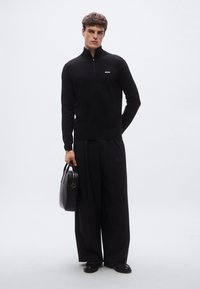 Svart pullover med glidelås i halsen og tettsittende design, med en liten hvit logo på brystet. Kombinert med vide svarte bukser og en svart veske.