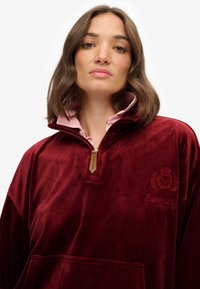 Sudadera burdeos de terciopelo con cuello con cremallera, con un interior de cuello rosa, bolsillo delantero y detalle de logo bordado.
