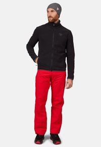 Schwarze Fleecejacke mit Reißverschluss und Logo, kombiniert mit leuchtend roten wasserdichten Hosen. Das Model trägt schwarze Schuhe und eine graue Mütze.