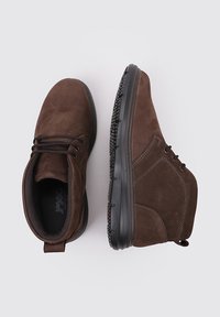Botas de tobillo de ante marrón con suelas de goma negras, con cordones en la parte delantera y detalles de costura, mostradas desde vistas superior y lateral sobre un fondo blanco.