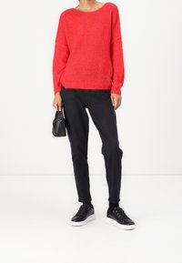 Roter, flauschiger Strickpullover mit weitem Ausschnitt, kombiniert mit schwarzen Slim-Fit-Jeans und schwarzen Sneakern. Das Model hält eine kleine schwarze Handtasche.