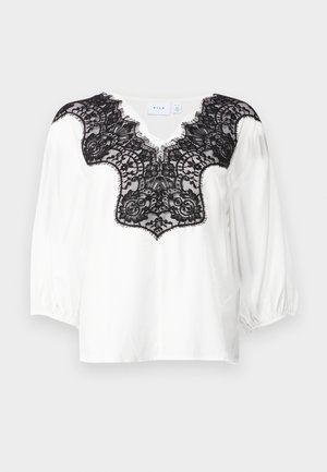 Balta bluzė su juoda nėriniuota apykakle, su V formos iškirpte ir pūstomis rankovėmis. Lygi medžiaga su sudėtingu nėriniuotu raštu.