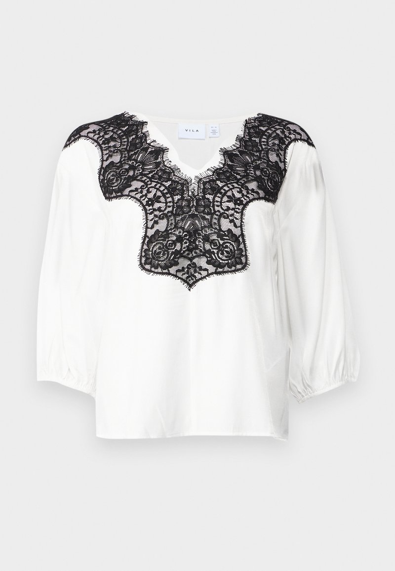 Blouse blanche avec des détails en dentelle noire au niveau du col, présentant une découpe en V et des manches bouffantes. Tissu lisse avec un design en dentelle complexe.