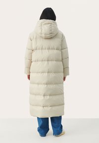 Lange beige pufferjas met een capuchon, met horizontale gestikte secties, gecombineerd met blauwe spijkerbroeken en beige sneakers.
